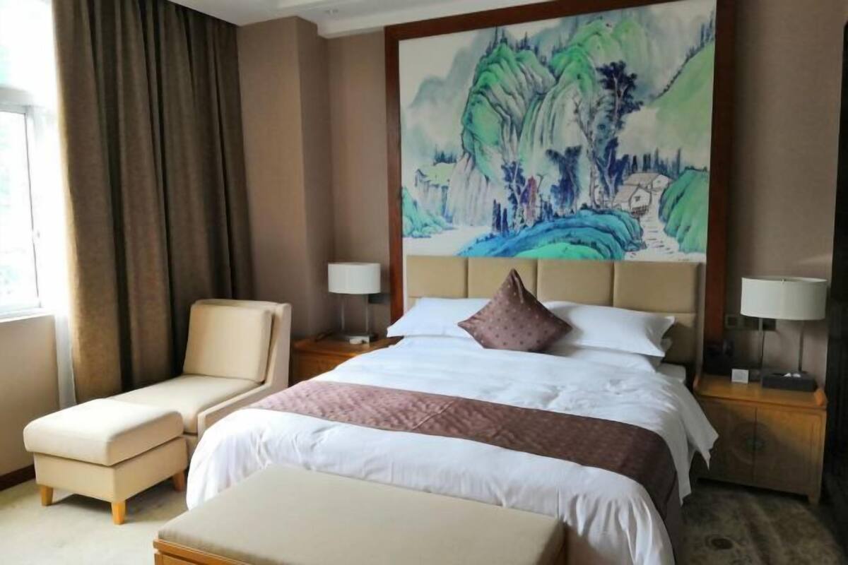 Een hotelkamer met een groot bed, een zithoek met een fauteuil, een nachtkastje met een lamp en een schilderij aan de muur.