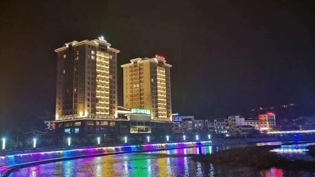 A high-rise hotel with multiple floors and balconies, a bridge with colorful lights, and a body of water in the foreground.