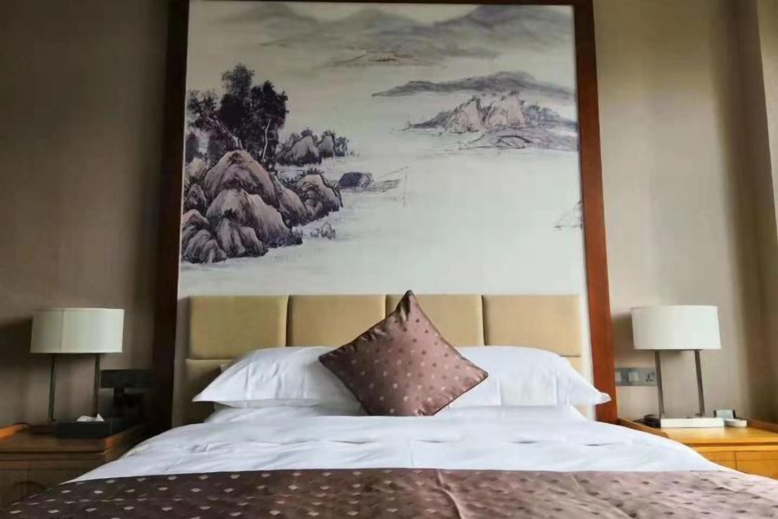 A hotel room with a large bed, a headboard, two bedside tables with lamps, and a large painting on the wall.