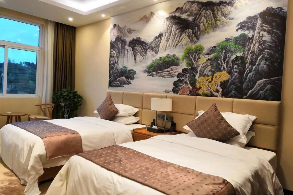 Een hotelkamer met twee bedden, een groot schilderij van een berglandschap, een raam en een nachtkastje met een lamp.