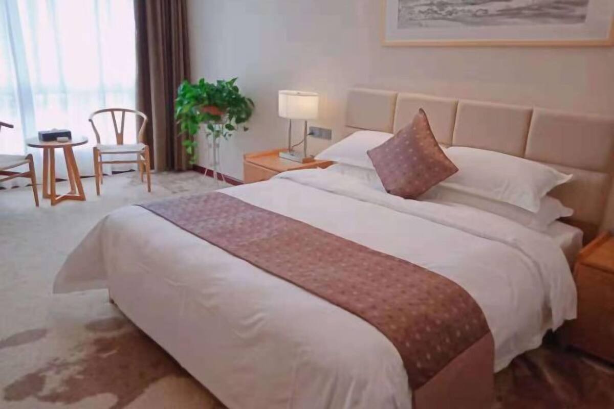 Een hotelkamer met een bed, nachtkastje, lamp, stoel en een plant.