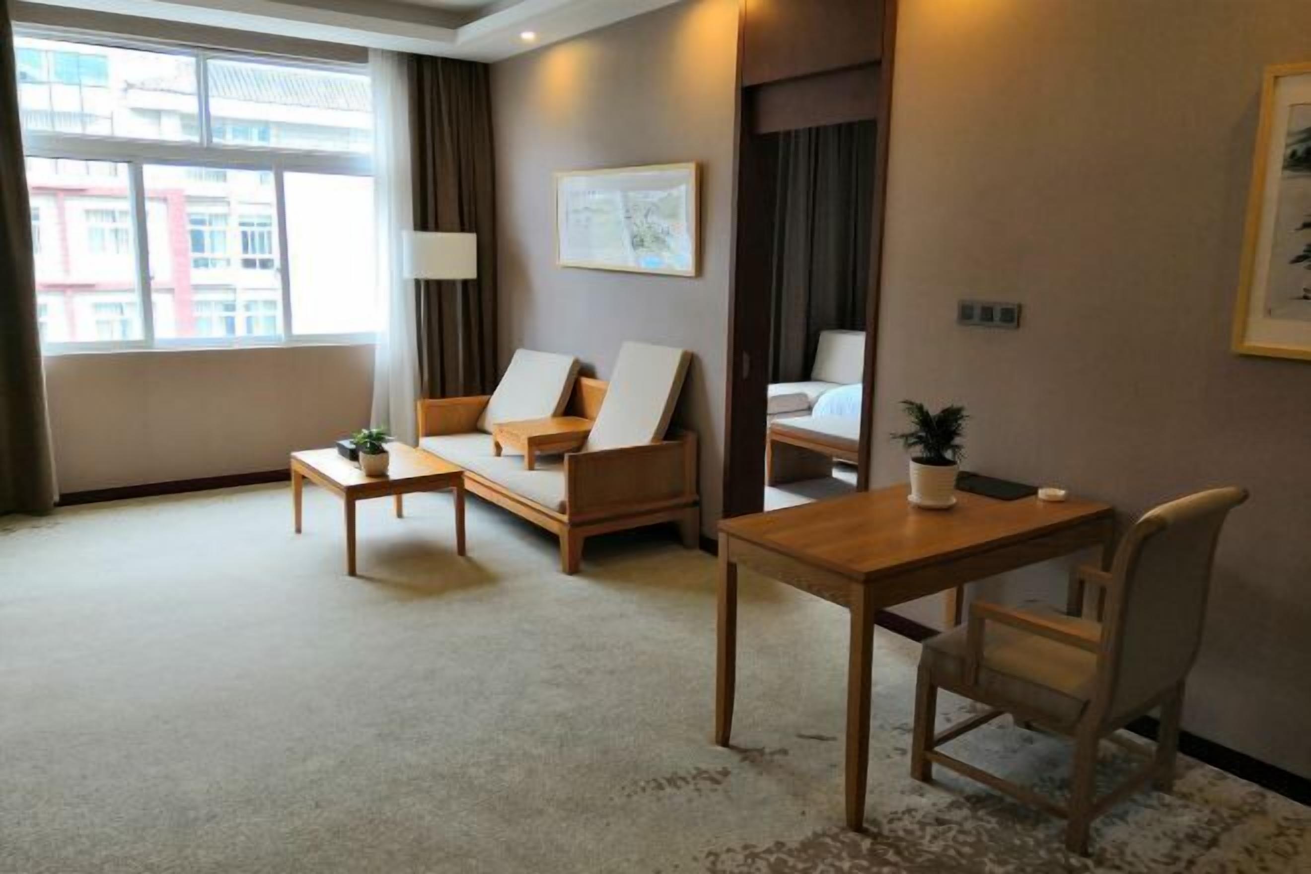 A hotel room with a sofa, a small table, a desk, and a chair. There is a window with curtains, a framed picture on the wall, and a potted plant on the desk.