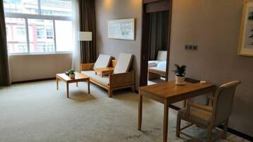 Une chambre d’hôtel avec un canapé, une petite table, un bureau et une chaise. Il y a une fenêtre avec des rideaux, un tableau encadré au mur et une plante en pot sur le bureau.