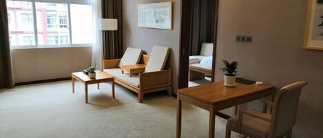 A hotel room with a sofa, a small table, a desk, and a chair. There is a window with curtains, a framed picture on the wall, and a potted plant on the desk.