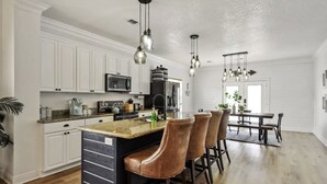 Una cocina moderna con armarios blancos, una isla central con taburetes de bar, electrodomésticos de acero inoxidable e iluminación colgante.