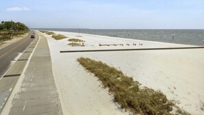 Una carretera costera con acera, zonas verdes y una playa al fondo.