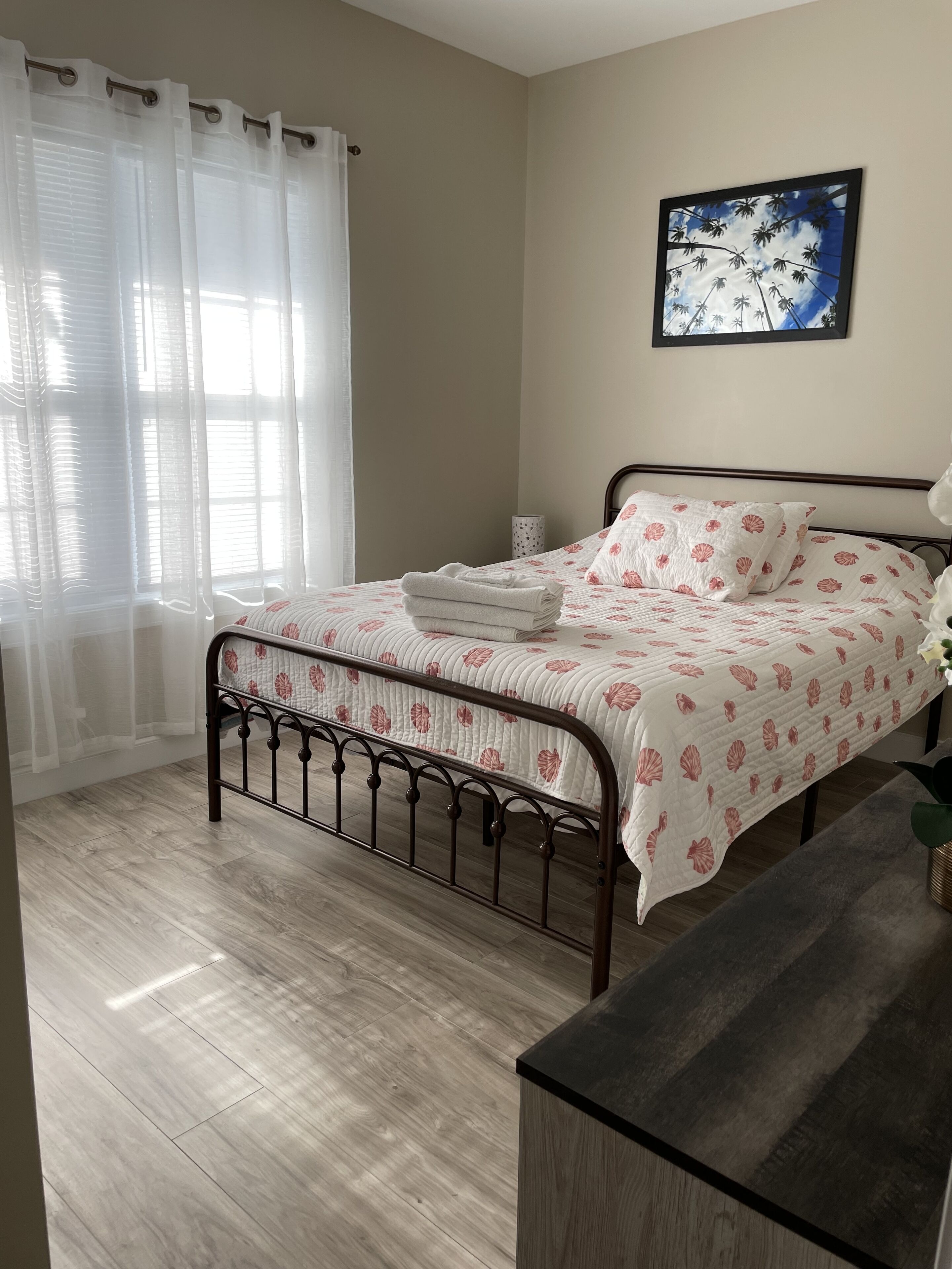 A bedroom with a bed, a bedside table, a framed picture on the wall, and a window with sheer curtains.