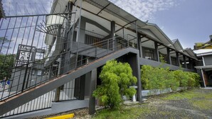 A modern building with a staircase leading to a covered area, surrounded by greenery and a gravel ground. - Coati Arenal Lodge (La Fortuna)