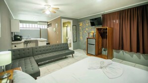 A hotel room with a bed, a sofa, a desk with a computer, a TV, and a bathroom visible through an open door. - Coati Arenal Lodge (La Fortuna)