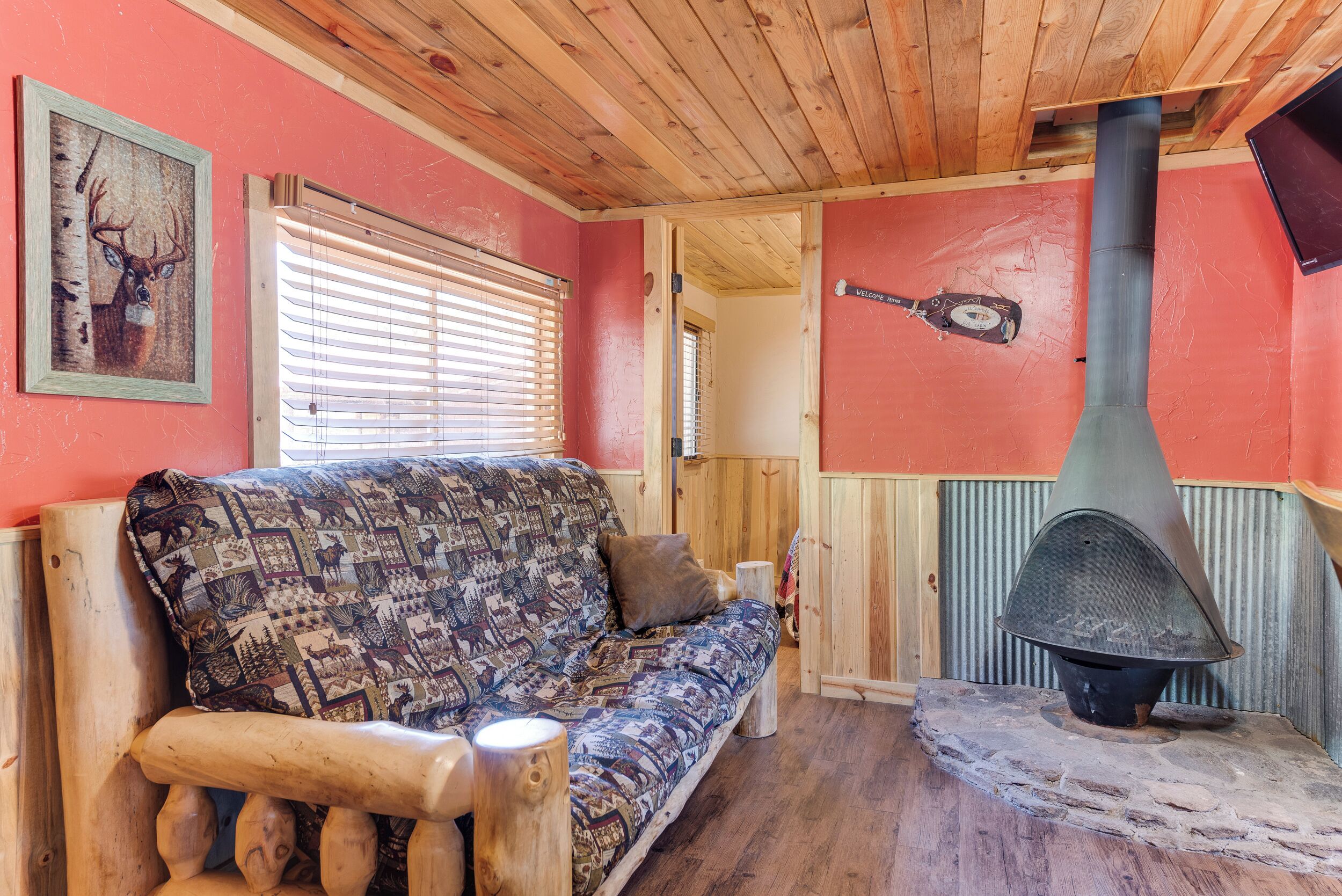 A cozy cabin interior with a wooden ceiling, a patterned sofa, a stone fireplace, and a framed deer painting.