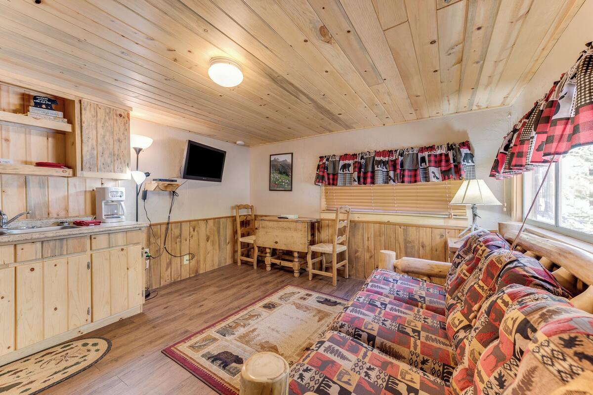 Ein gemütliches Blockhaus-Interieur mit Holzwänden und -böden, einer Couch mit gemusterten Kissen, einem Esstisch mit Stühlen und einer Küchenzeile mit Kaffeemaschine und Schränken.