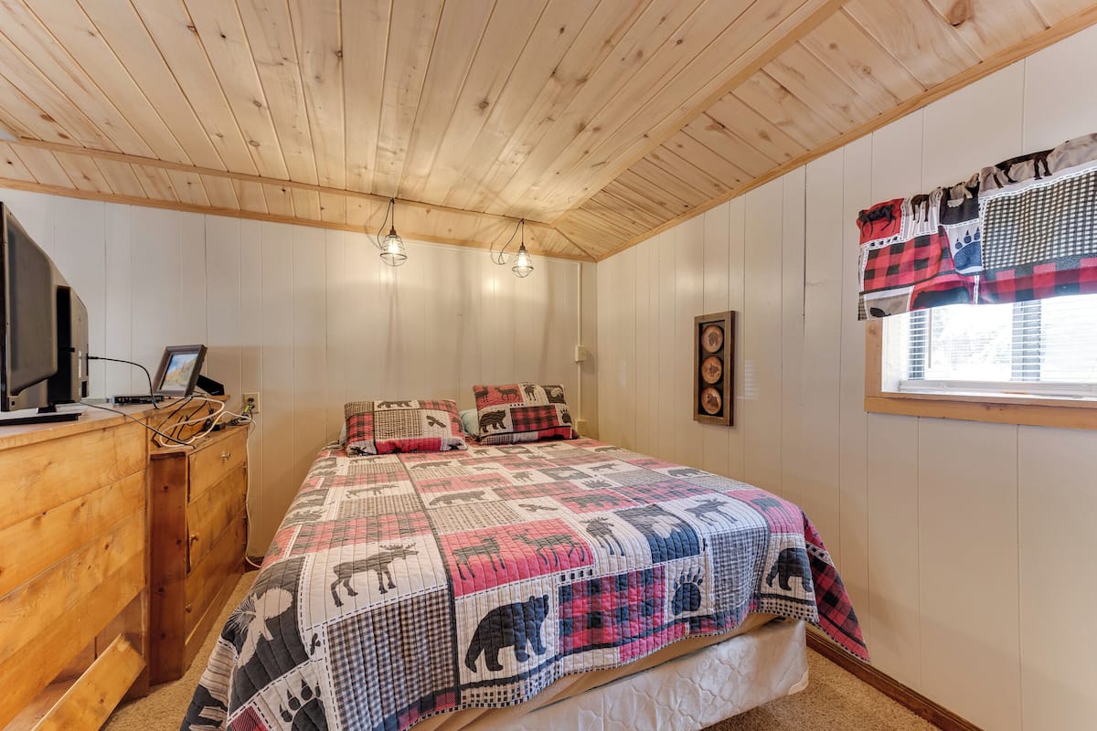 Ein Schlafzimmer mit Holzboden, einem Bett mit gesteppter Decke, einem Schminktisch, einem Fernseher und einem Fenster mit Vorhängen.