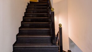 Una escalera con escalones oscuros y barandilla, que conduce a un rellano con una lámpara y un cuadro enmarco.