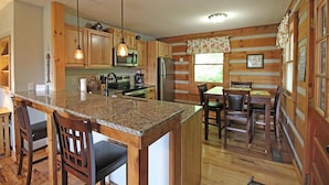 A kitchen with wooden cabinets, granite countertops, a central island with bar stools, and a dining area with a table and chairs. - True Log Cabin (Bryson City)