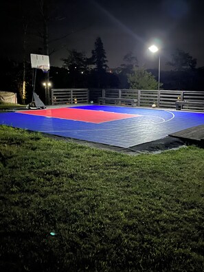 Un terrain de basket en plein air, éclairé la nuit, avec une surface de jeu bleue et rouge, entouré d’une clôture en bois, et un lampadaire en arrière-plan.