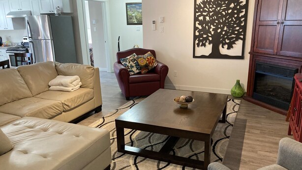 Ein Wohnzimmer mit einem beigen Sofa, einem roten Sessel, einem Couchtisch mit einer Snackschale, einem Kamin und einer Küche im Hintergrund.