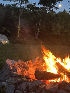 Un feu de camp avec des bûches et des rochers, entouré d’une zone herbeuse avec des arbres et une formation rocheuse en arrière-plan.