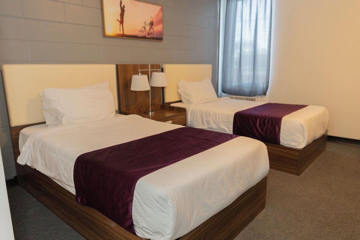 Dos camas individuales con cabeceros de madera y ropa de cama blanca, una mesita de noche con dos lámparas y un cuadro en la pared.