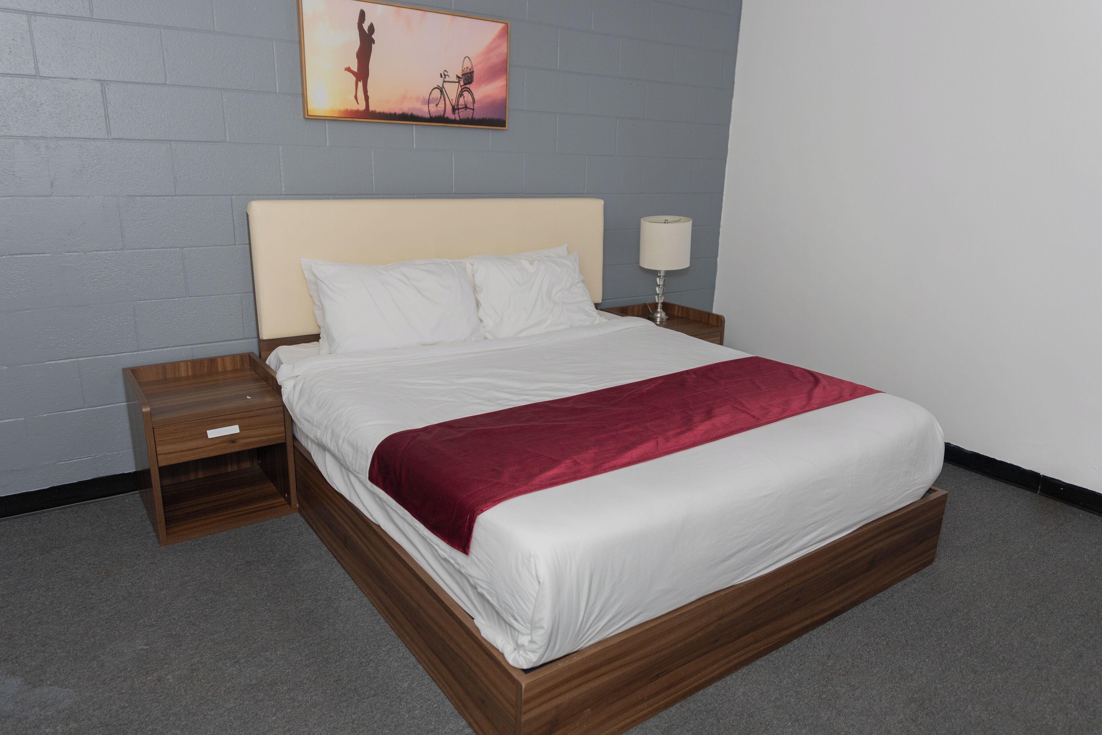 A neatly made bed with a wooden frame, a white headboard, and a red throw blanket. There is a nightstand with a lamp beside the bed.