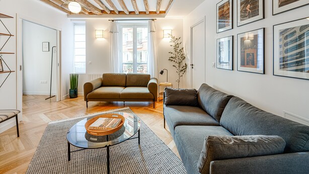 A modern living room with a wooden ceiling, a grey sofa, a round glass coffee table, and framed artwork on the walls. - Appartement Grenelle - Welkeys (Paris)