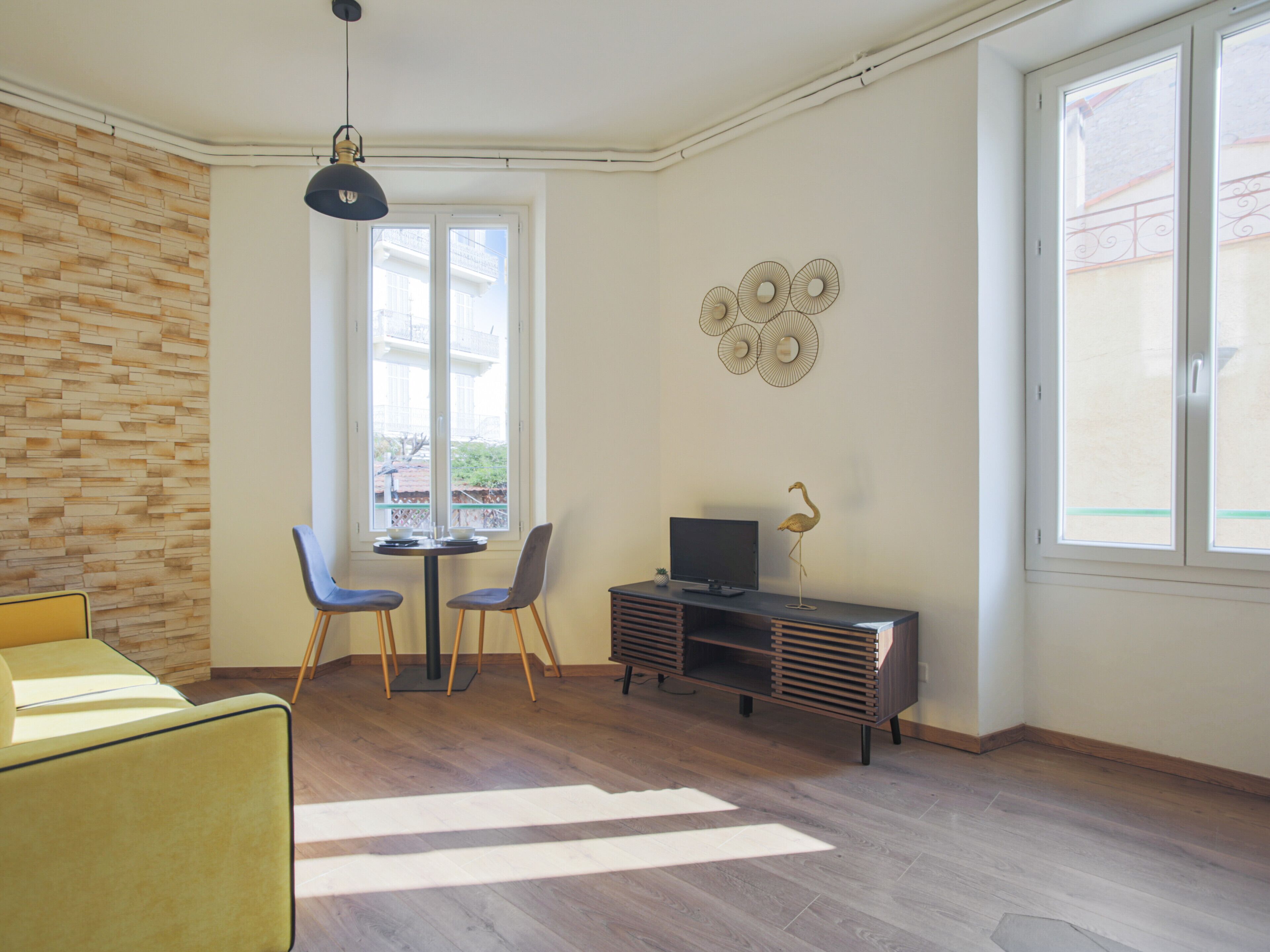 A modern living room with a wooden floor, a yellow sofa, a dining table with two chairs, a TV on a wooden cabinet, and a large window with a view of buildings.