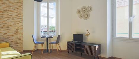 A modern living room with a wooden floor, a yellow sofa, a dining table with two chairs, a TV on a wooden cabinet, and a large window with a view of buildings.