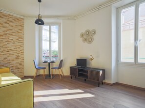 Uma sala de estar moderna com piso de madeira, um sofá amarelo, uma mesa de jantar com duas cadeiras, uma TV em um móvel de madeira e uma janela grande com vista para edifícios.