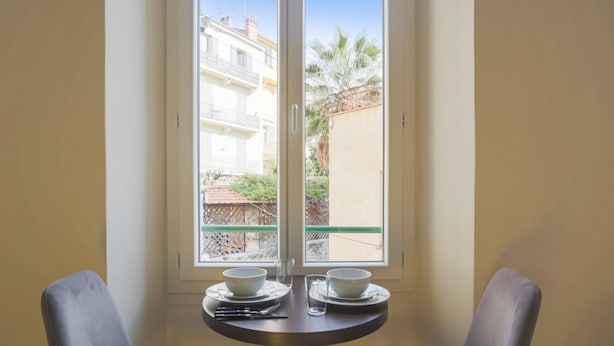 A room with a table and two chairs, a window with a view of buildings and a fence. - Studio Nid Doré - Welkeys (Cannes)