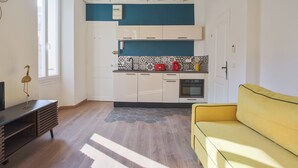A compact kitchen with white cabinets, a blue accent wall, a yellow sofa, and a wooden floor. - Studio Nid Doré - Welkeys (Cannes)