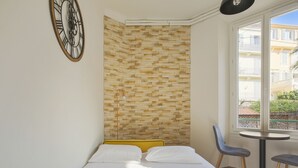 A hotel room with a bed, a clock on the wall, a window with a view of buildings, and a small table with chairs. - Studio Nid Doré - Welkeys (Cannes)