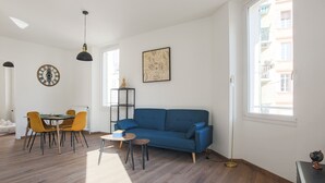 Ein modernes Wohnzimmer mit einem blauen Sofa, Holzboden, einem Esstisch mit gelben Stühlen und einer schwarzen Pendelleuchte.