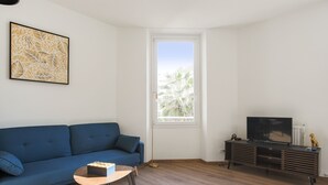 Ein Wohnzimmer mit blauem Sofa, einem Couchtisch, einem Fernseher auf einem Holzschrank und einem Fenster mit Blick auf Palmen.