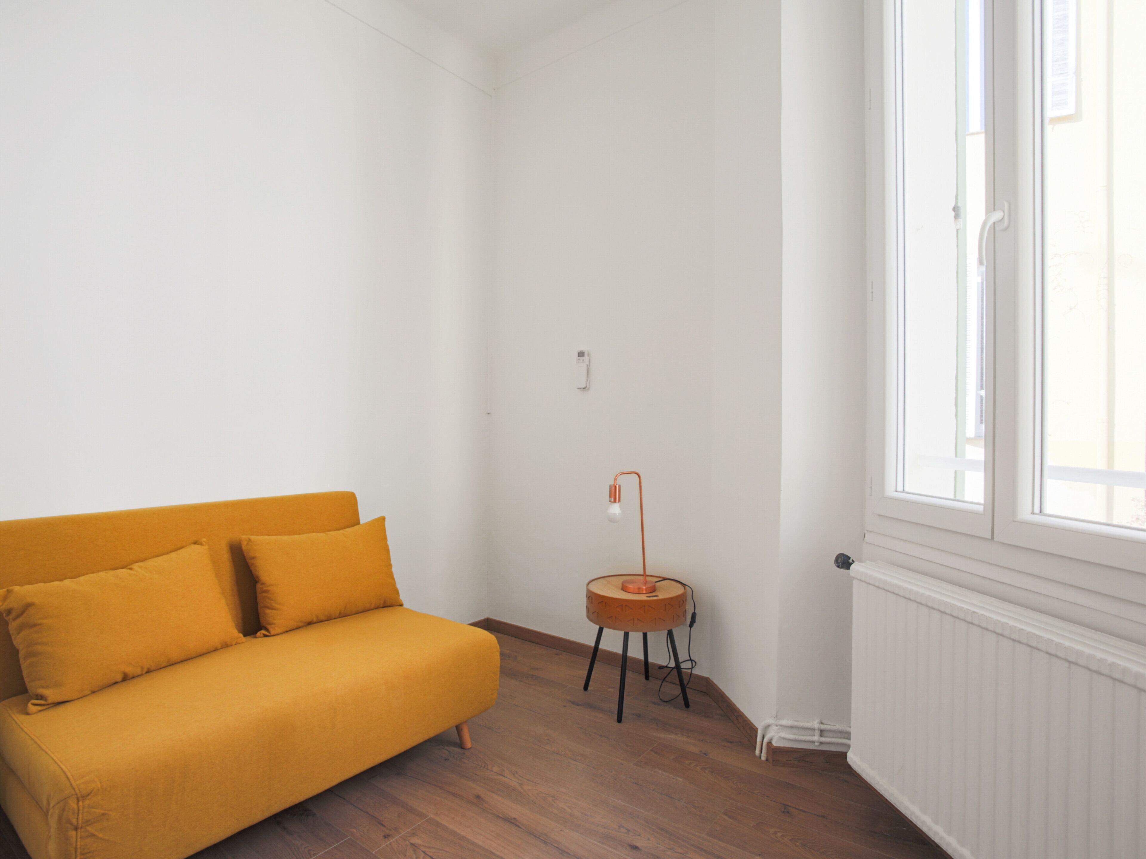 Um quarto claro, com mobiliário simples, piso de madeira, um sofá amarelo-mostarda, uma pequena mesa de madeira com uma luminária e uma janela com molduras brancas.