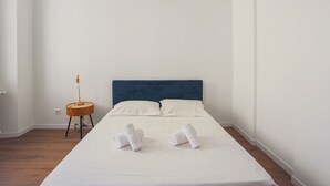 Ein ordentlich bezogenes Bett mit weißen Leinen und zwei aufgerollten Handtüchern, ein kleiner runder Tisch mit einer Lampe und ein blaues Kopfstück.
