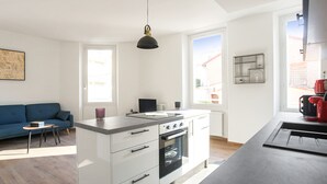 Eine moderne Küche mit weißen Schränken, einer schwarzen Arbeitsplatte und einem eingebauten Ofen. Im Wohnbereich befindet sich eine blaue Couch und ein kleiner Tisch.