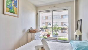 Hotellihuone, jossa on sänky, ikkuna, josta on näkymä rakennuksiin, ja parveke, sekä tuoli, pöytä ja taulu seinällä.