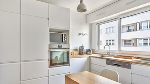 Moderni keittiö, jossa on valkoiset kaapit, sisäänrakennettu uuni, mikroaaltouuni ja astianpesukone, puinen pöytä ja ikkuna, josta on näkymä rakennukseen.