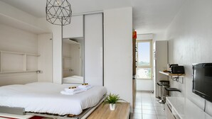 A modern hotel room with a large bed, a small table with a plant, a TV, and a kitchenette. - Studio Doris - Welkeys (Montpellier)