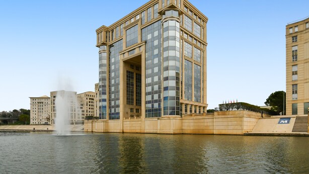 A modern building with a large glass facade, a fountain in front, and a clear blue sky. - Studio Doris - Welkeys (Montpellier)