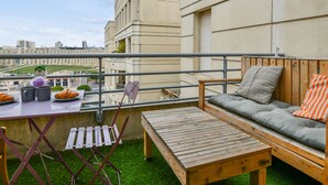 A balcony with a wooden table, a cushioned sofa, and a view of city buildings. - Studio Doris - Welkeys (Montpellier)