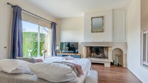 A bedroom with a large bed, a TV, a fireplace, and a view of greenery outside. - Appartement Amirauté - Welkeys (Tourgéville)