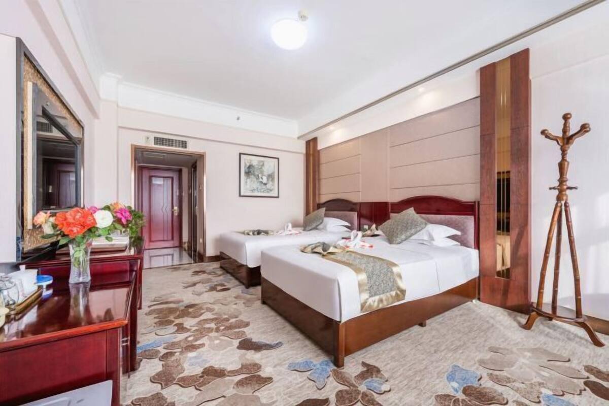 A hotel room with two beds, a desk, a mirror, a wooden headboard, a standing lamp, and a patterned carpet.