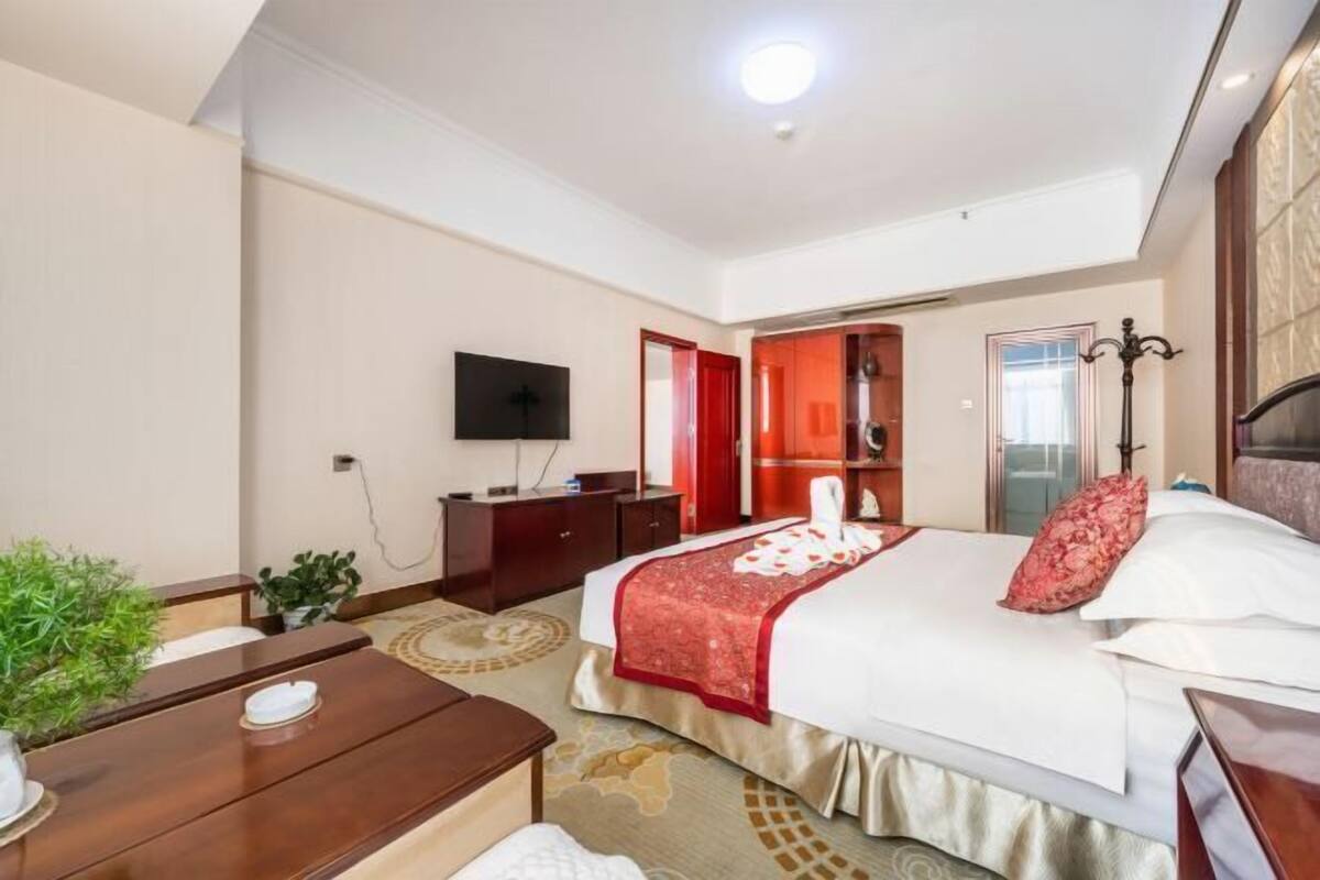 A hotel room with a bed, a TV, a desk, a chair, a plant, and a red door.