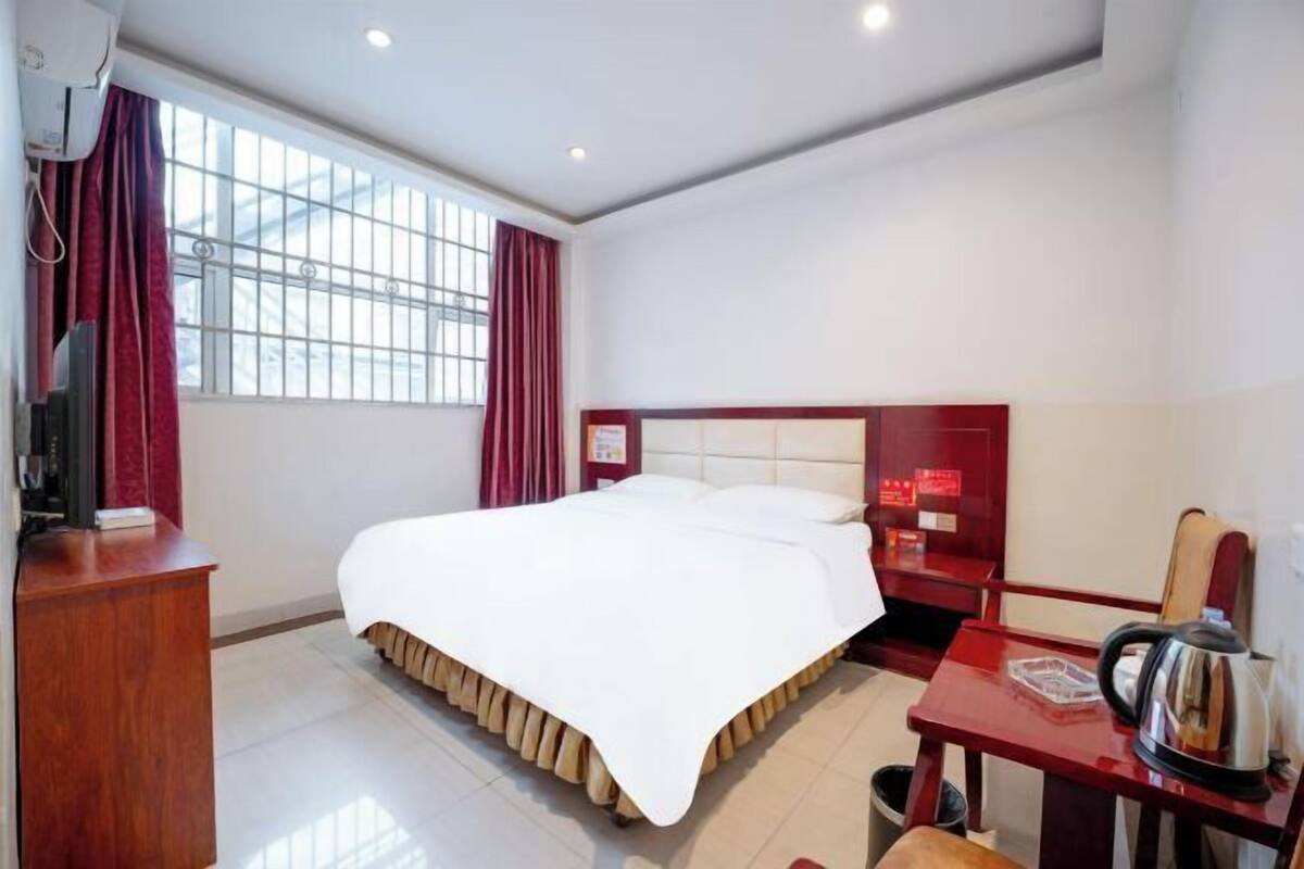 A hotel room with a large bed, a wooden desk, a kettle, and a window with curtains.