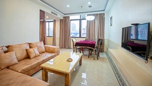 A living room with a leather sofa, a wooden coffee table, a dining table with chairs, a flat-screen TV mounted on the wall, and a window with curtains.