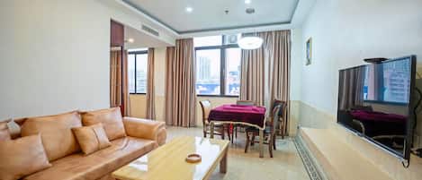 A living room with a leather sofa, a wooden coffee table, a dining table with chairs, a flat-screen TV mounted on the wall, and a window with curtains.