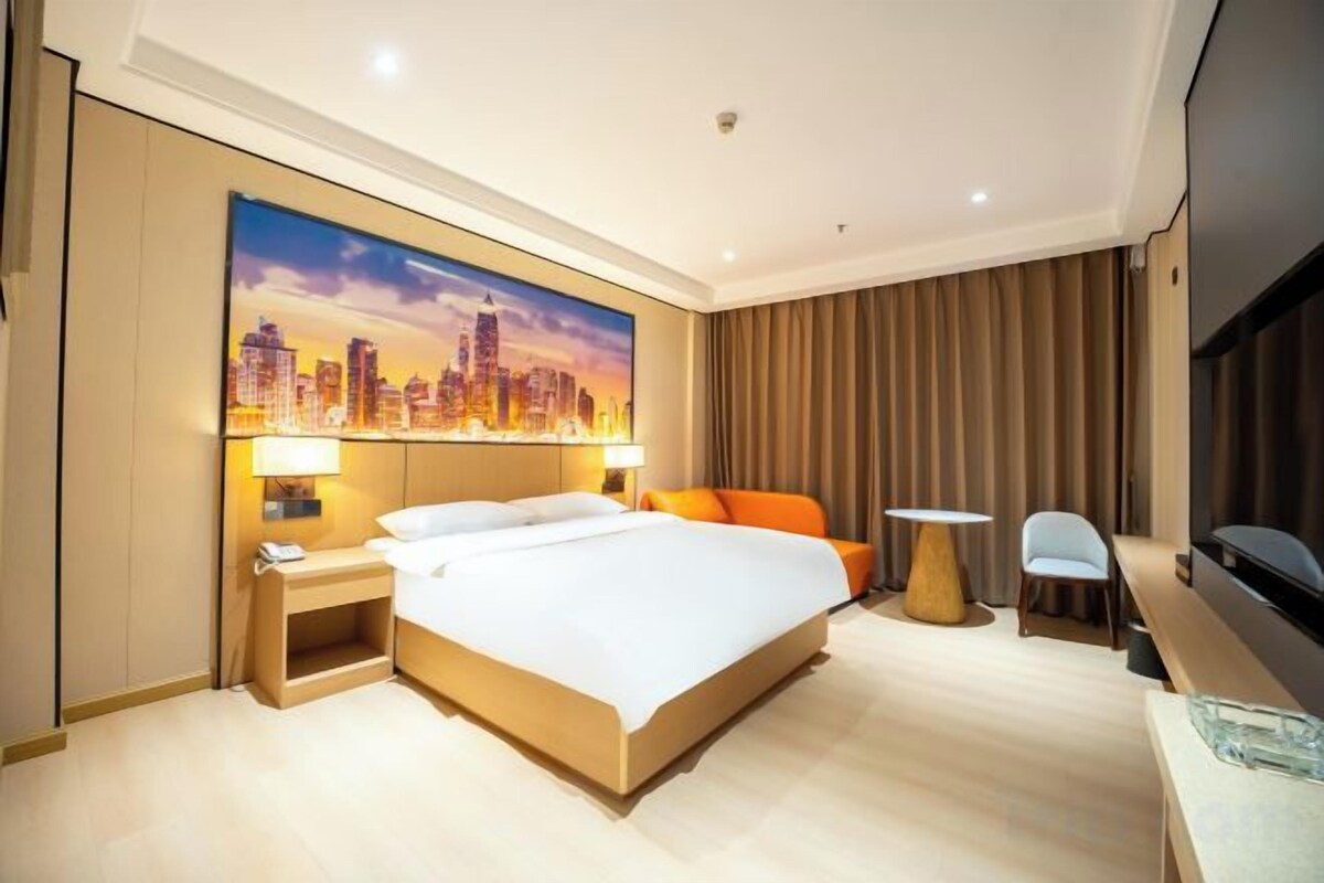 A hotel room with a large bed, a desk, a chair, a TV, and a cityscape painting on the wall.