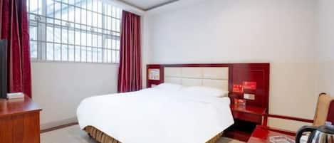 A hotel room with a large bed, a desk with a chair, a red headboard, and a window with curtains.