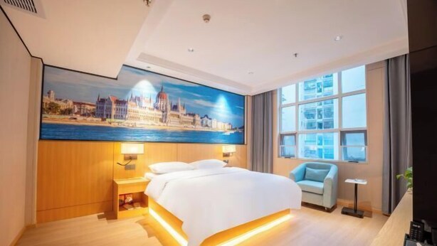 A hotel room with a bed, a desk, a chair, a window with curtains, and a cityscape painting on the wall. - Huifeng Hotel (Lechang)