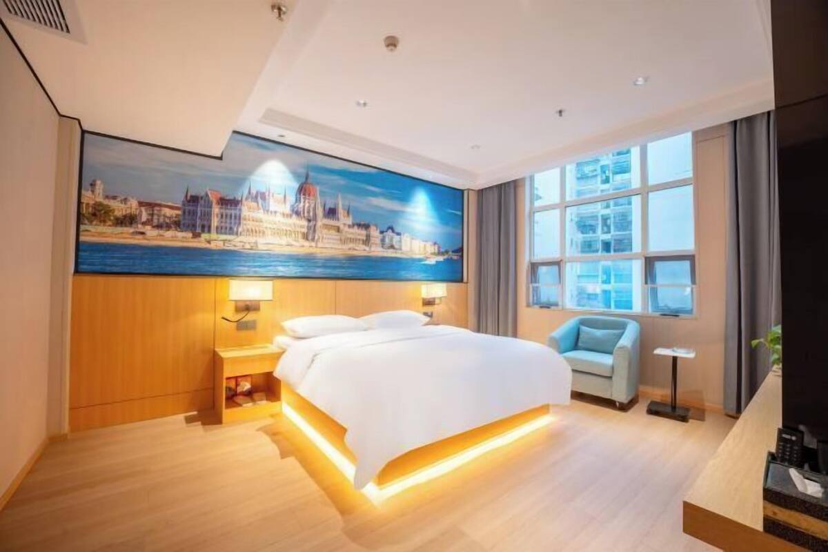A hotel room with a bed, a desk, a chair, a window with curtains, and a cityscape painting on the wall.